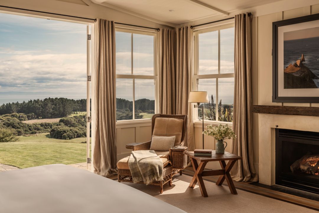 A hotel suite with a fireplace, windows, and neutral furnishings, including a plush bed, wicker chairs, and soft curtains.